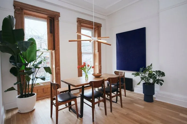 Deep blue canvas on a white dining-room wall between wooden window frames, with a slim wooden table, brown chairs, plants, and a wooden linear light.