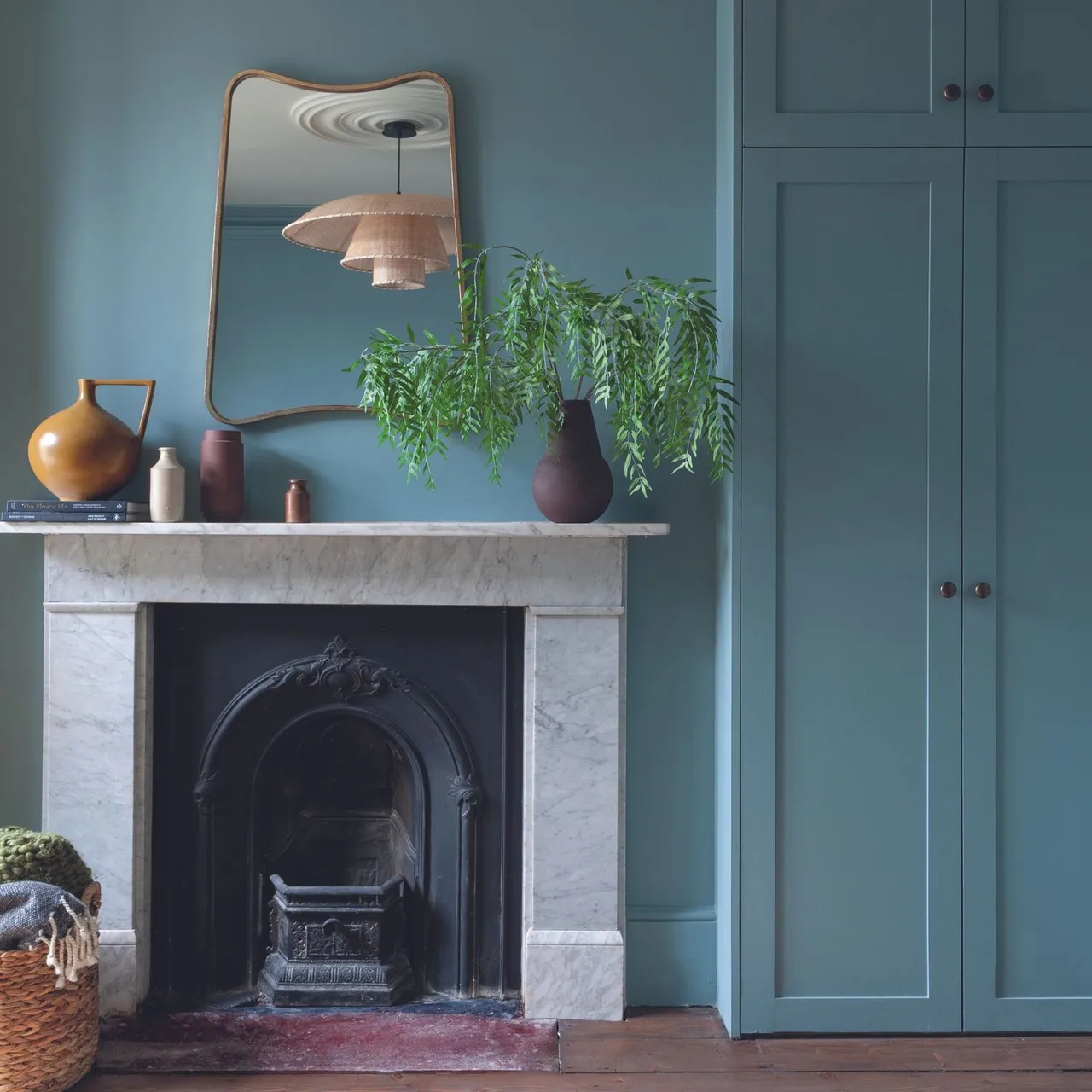 Pale veined fireplace against teal wall with small warm-colored vessels, tall mirror and leafy branch arrangement