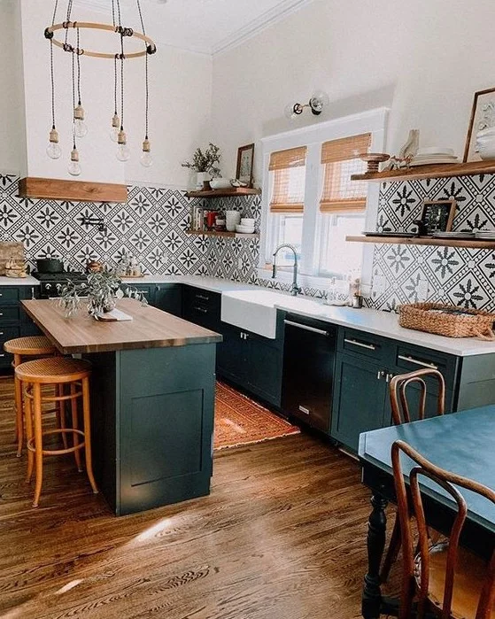 Deep green kitchen cabinets and an island stand below black-and-white geometric tiles beside wooden shelves, wood flooring and a white apron-front sink