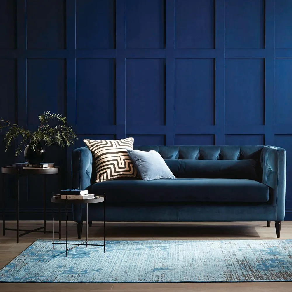 A blue sofa stands in front of darker blue paneling with a pale rug below.