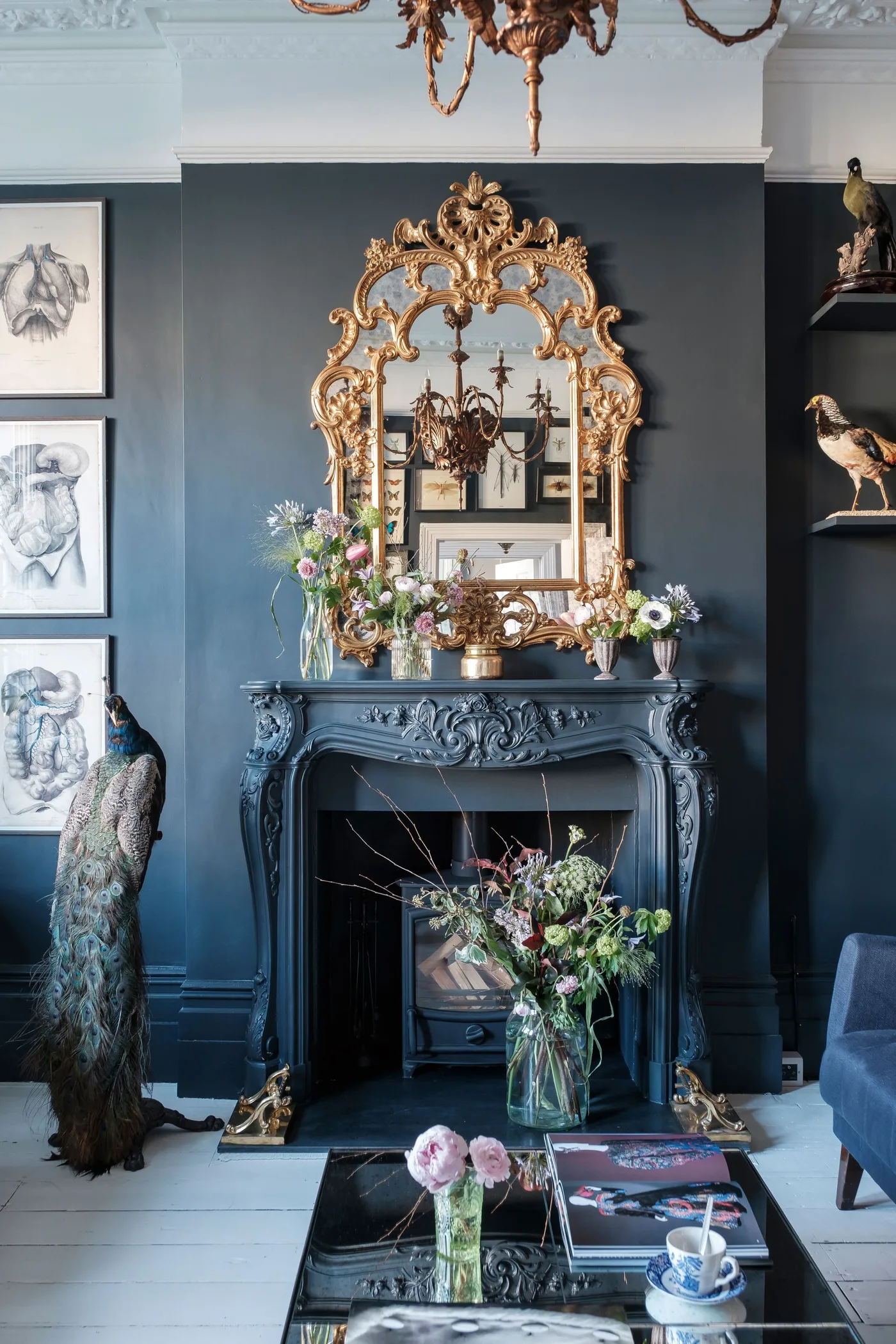 An ornate dark blue fireplace mantel sits beneath a large gold-framed mirror beside framed pictures and blue seating