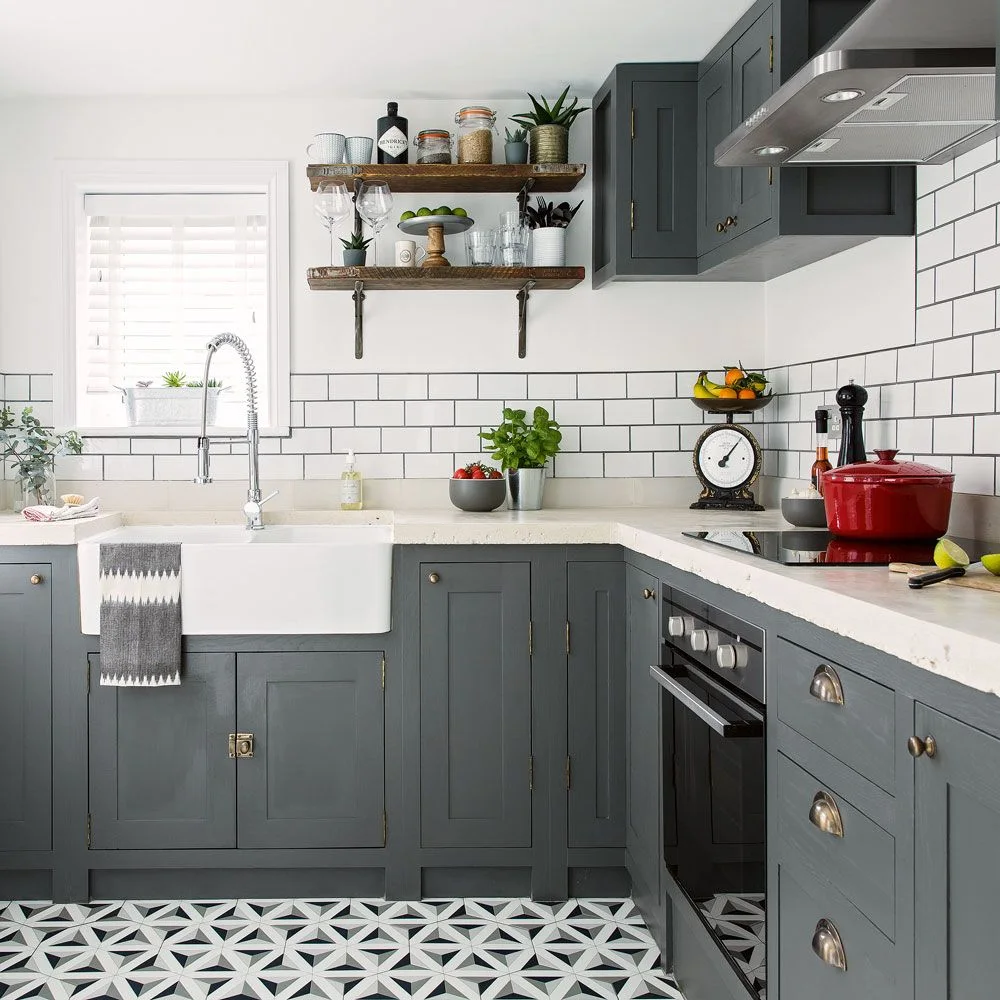 Dark gray paneled cabinets sit below white rectangular tiles beside a white apron-front sink, wooden shelves and patterned flooring