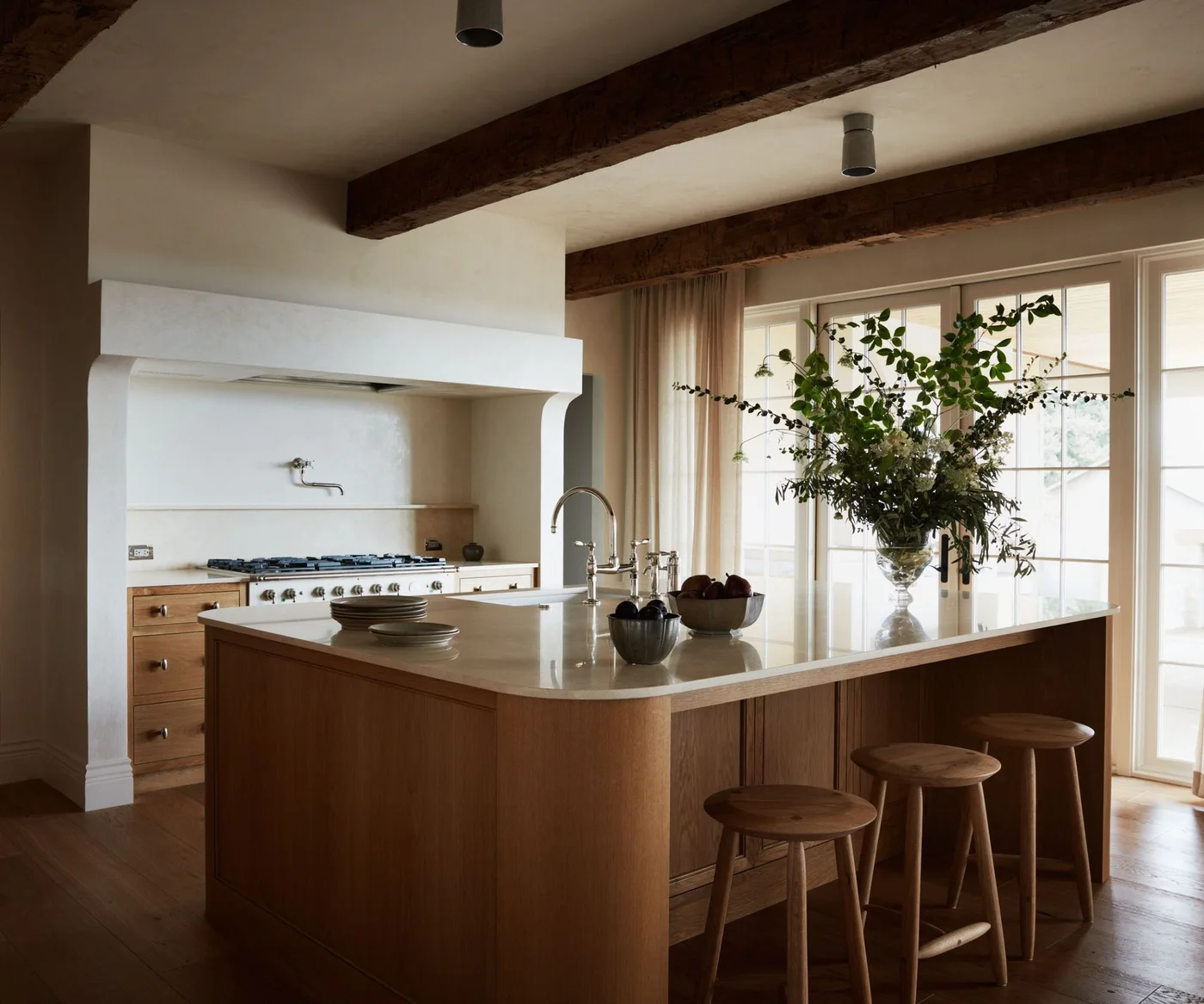 A wooden kitchen island has a rounded pale counter edge beside three wooden stools, a pale hood, dark ceiling beams and tall curtains