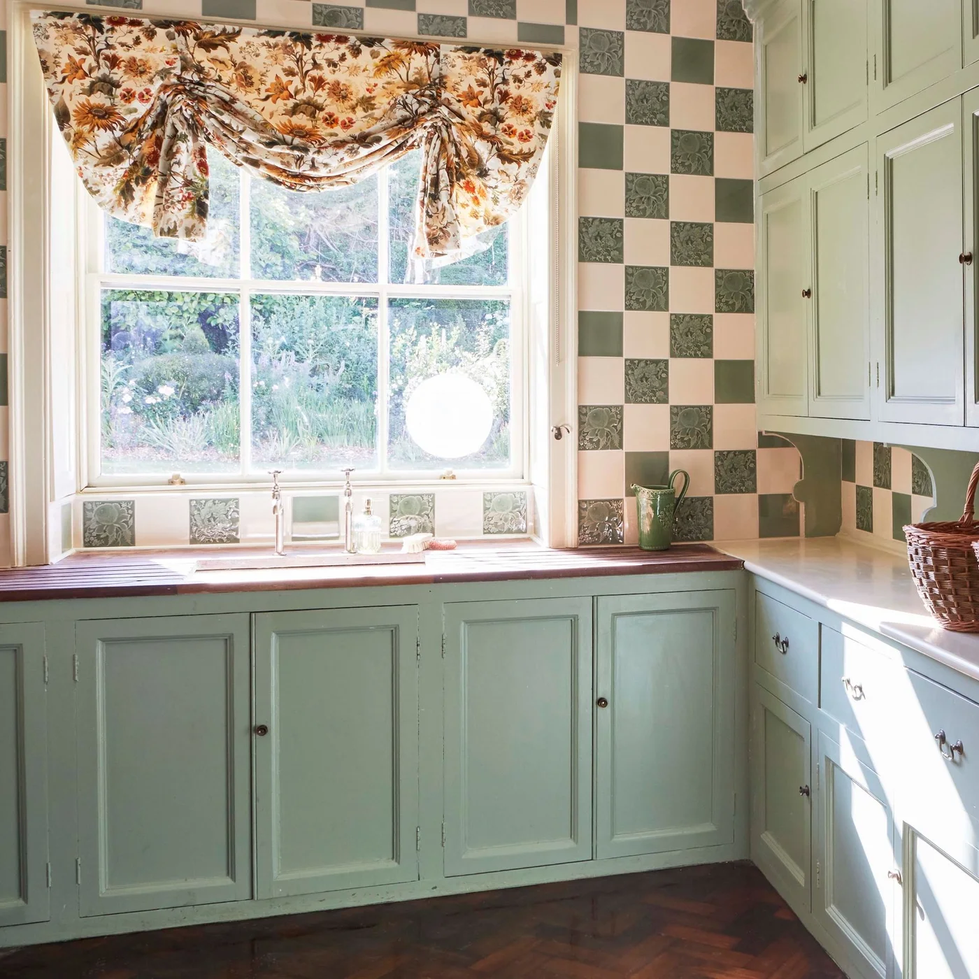 Pale green cabinets sit below green-and-pale checked wall tiles beside a window with a floral fabric treatment