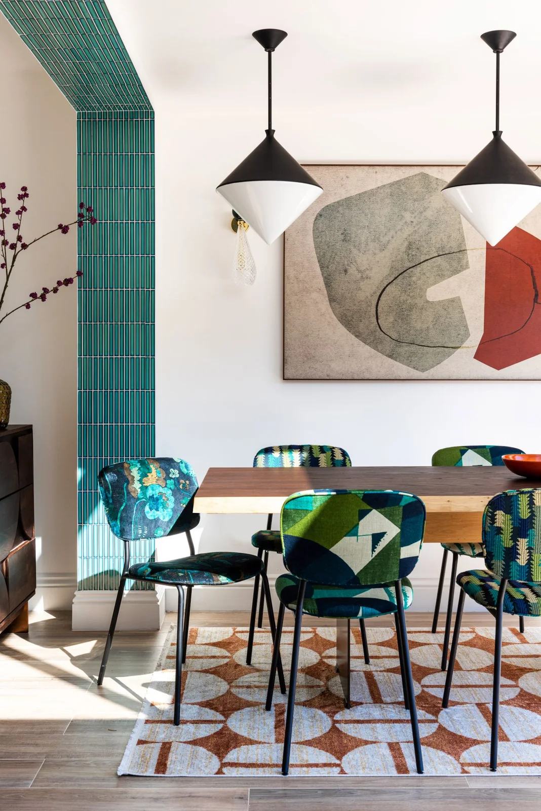 Teal and green patterned chairs surround a wooden table above a rust-and-white geometric rug beneath two black-and-white pendants