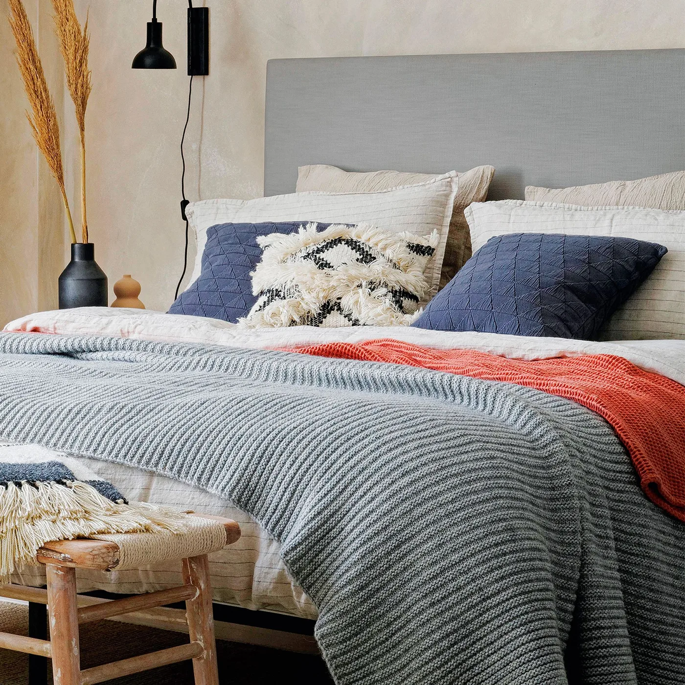 Quilted navy cushions on pale bedding with a gray upholstered headboard, knitted gray throw, and a woven bedside stool.