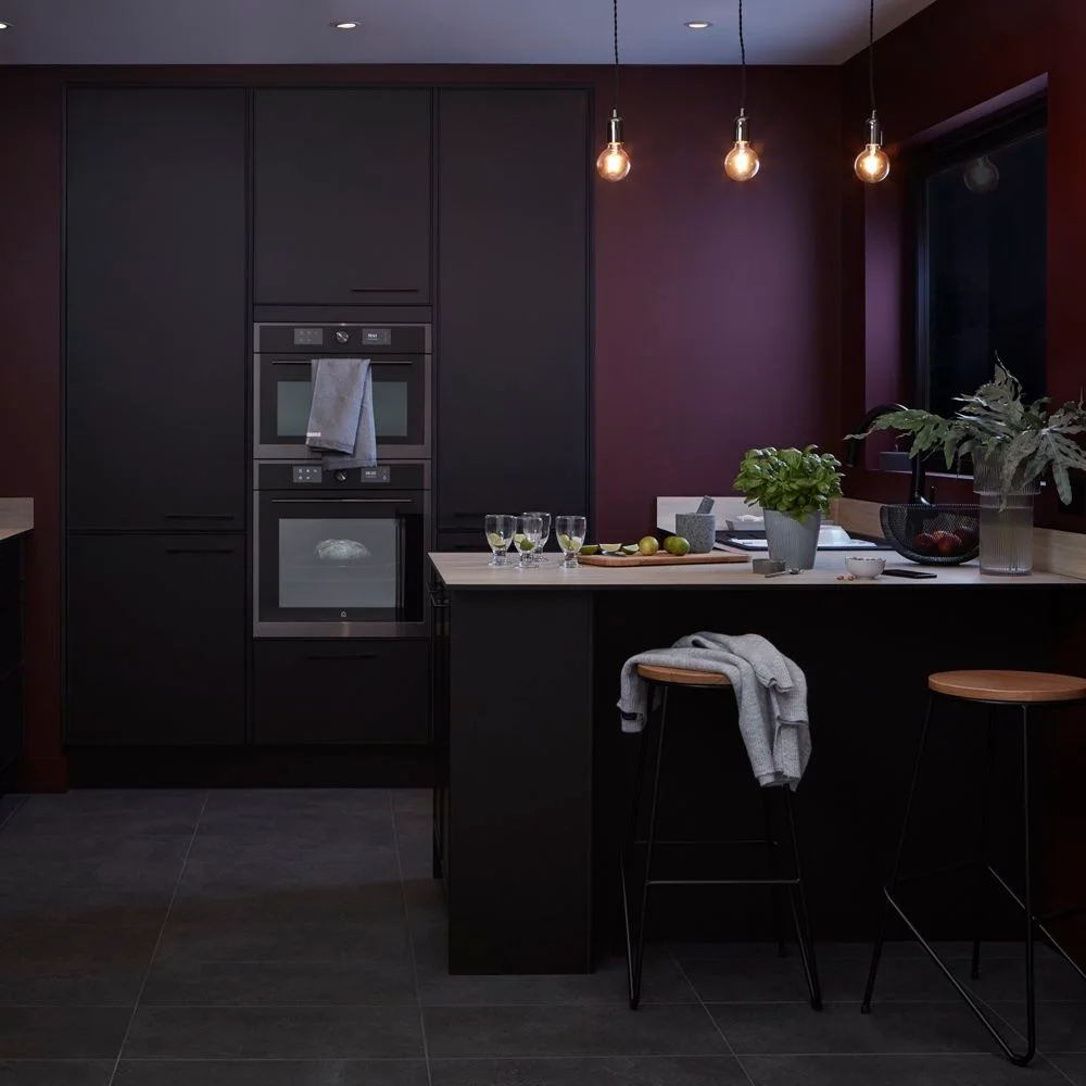 Dark kitchen cupboards and a peninsula sit beside a deep burgundy wall, three small hanging bulbs and a plain wood-topped stool