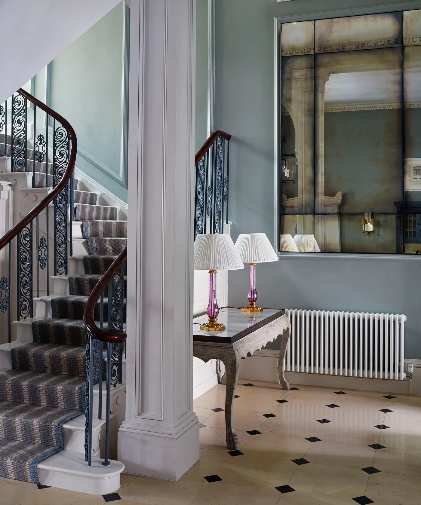 Narrow traditional console with two colored glass lamps and pale pleated shades beside a patterned stair runner and large mirror