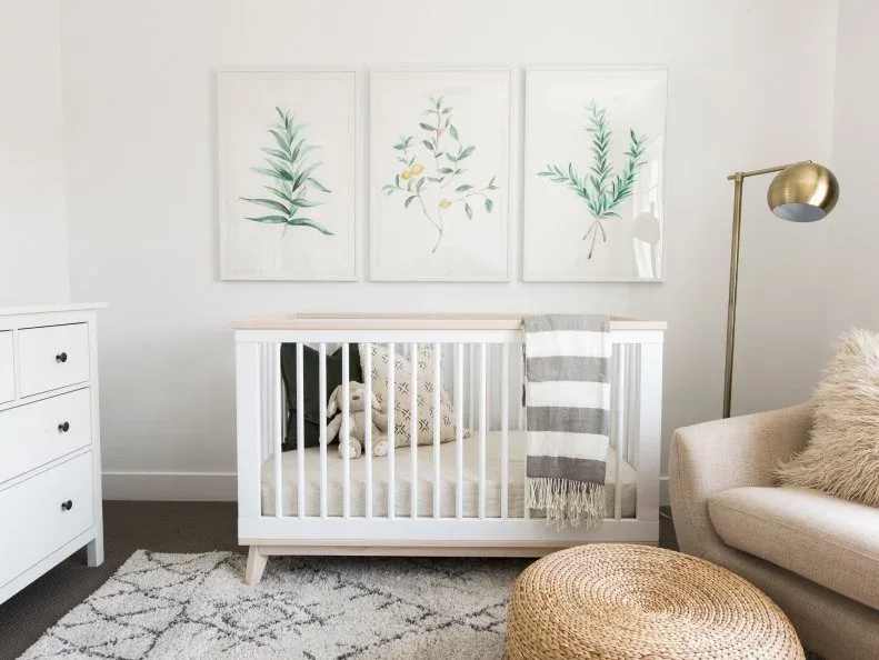 White crib beneath botanical pictures beside pale adult sofa, woven pouf, patterned rug and floor lamp.