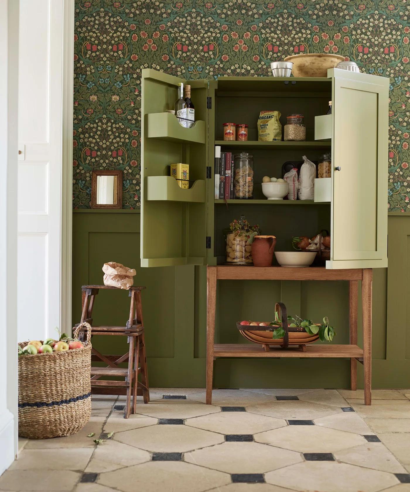 An open green pantry cabinet holds jars, books and food containers above a wooden lower table beside floral wallpaper and a pale patterned floor