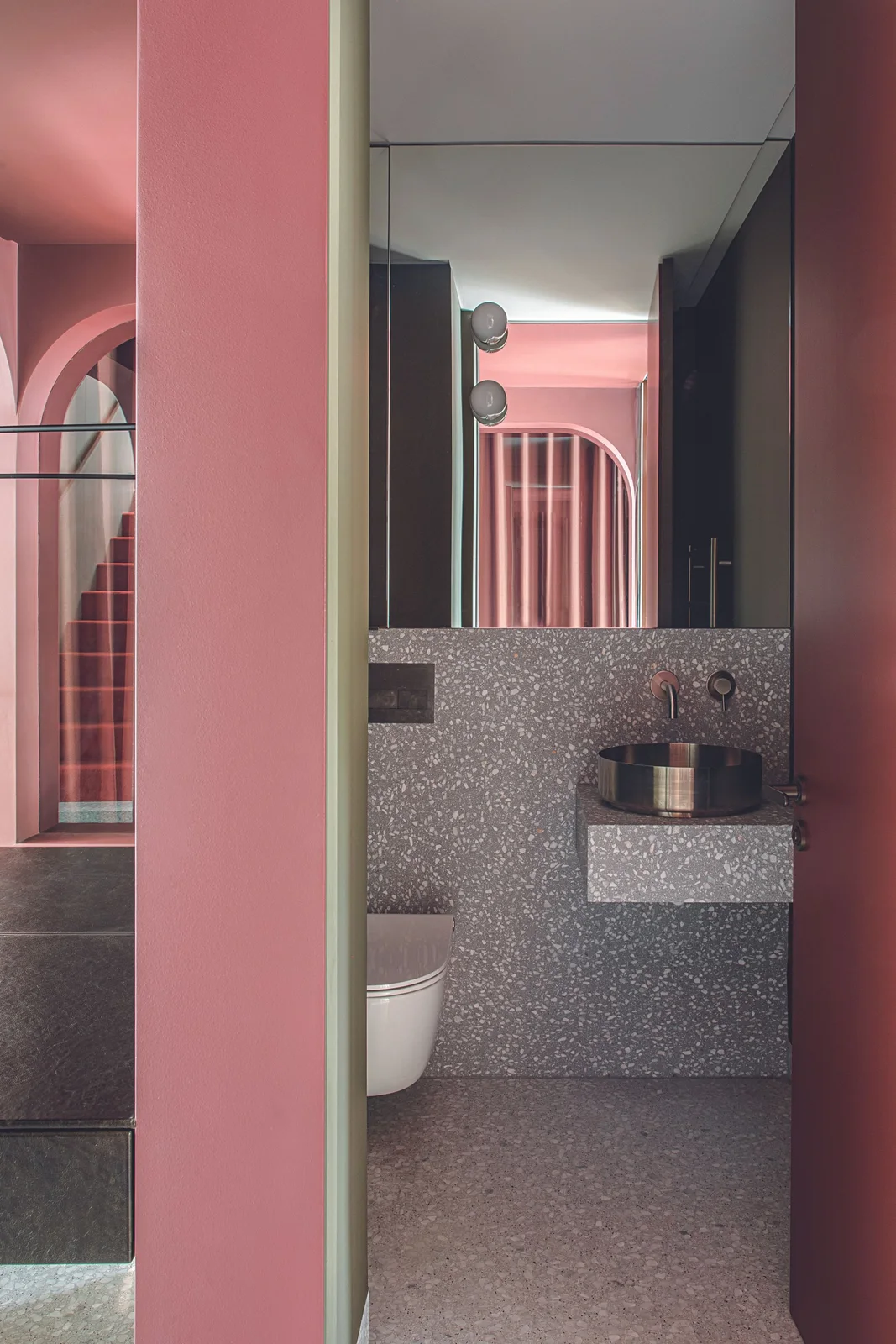 A small dark round basin sits on a gray speckled wall-mounted shelf beside pink architectural walls, tall mirror panels and a white toilet
