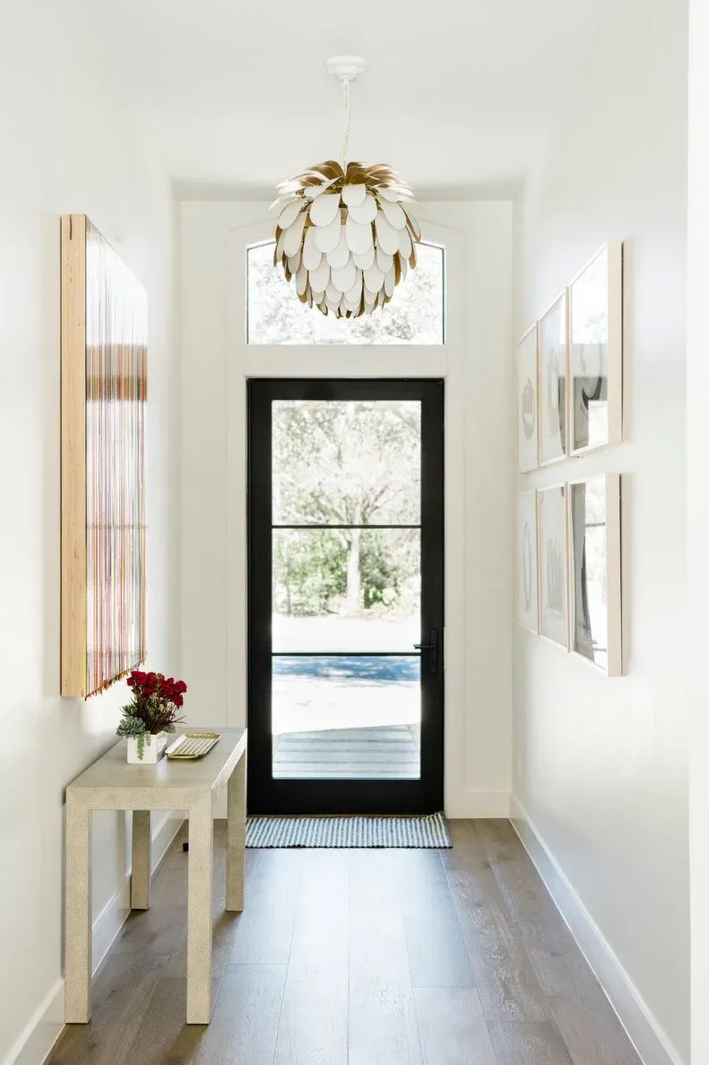 Narrow white entrance with black glazed door, small pale console, wall pictures and layered white pendant.