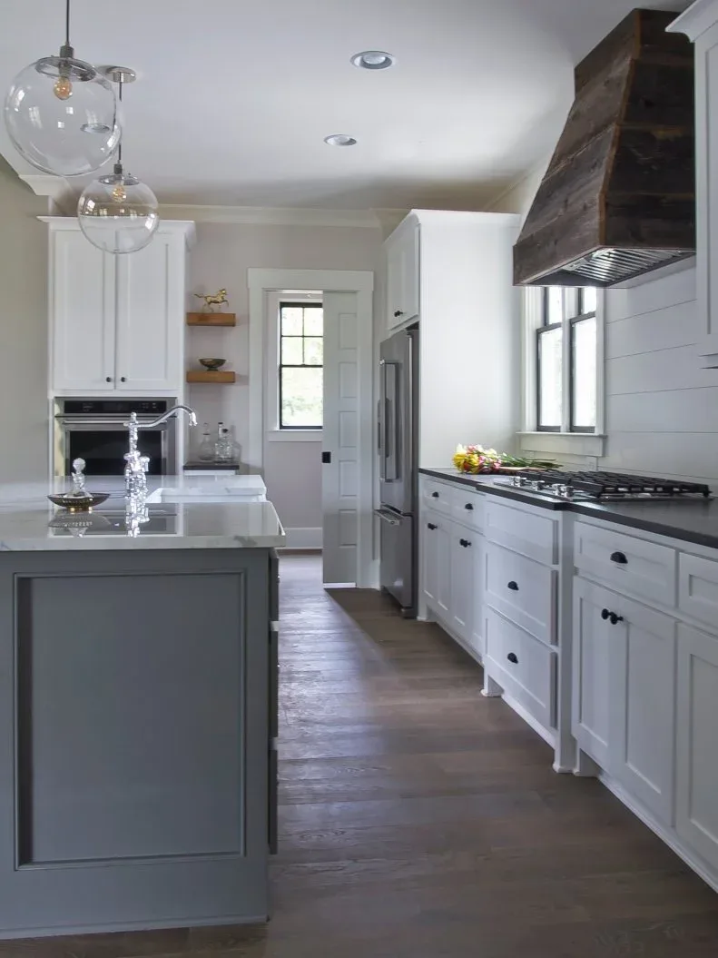 White kitchen with gray island, dark wood range-hood surround, clear globe pendants and wood floor