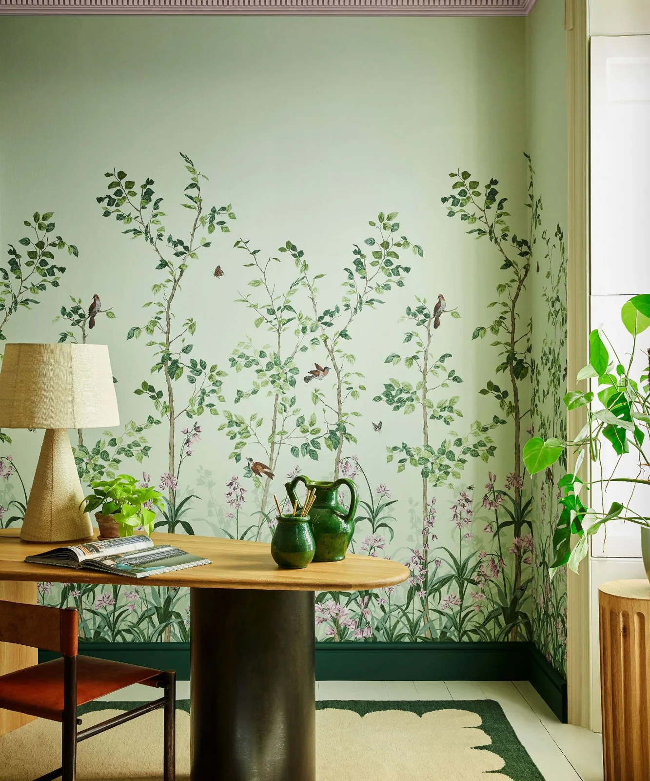 Pale green leafy wallpaper sits behind a rounded wooden desk with a shaded lamp, orange chair and green pitchers
