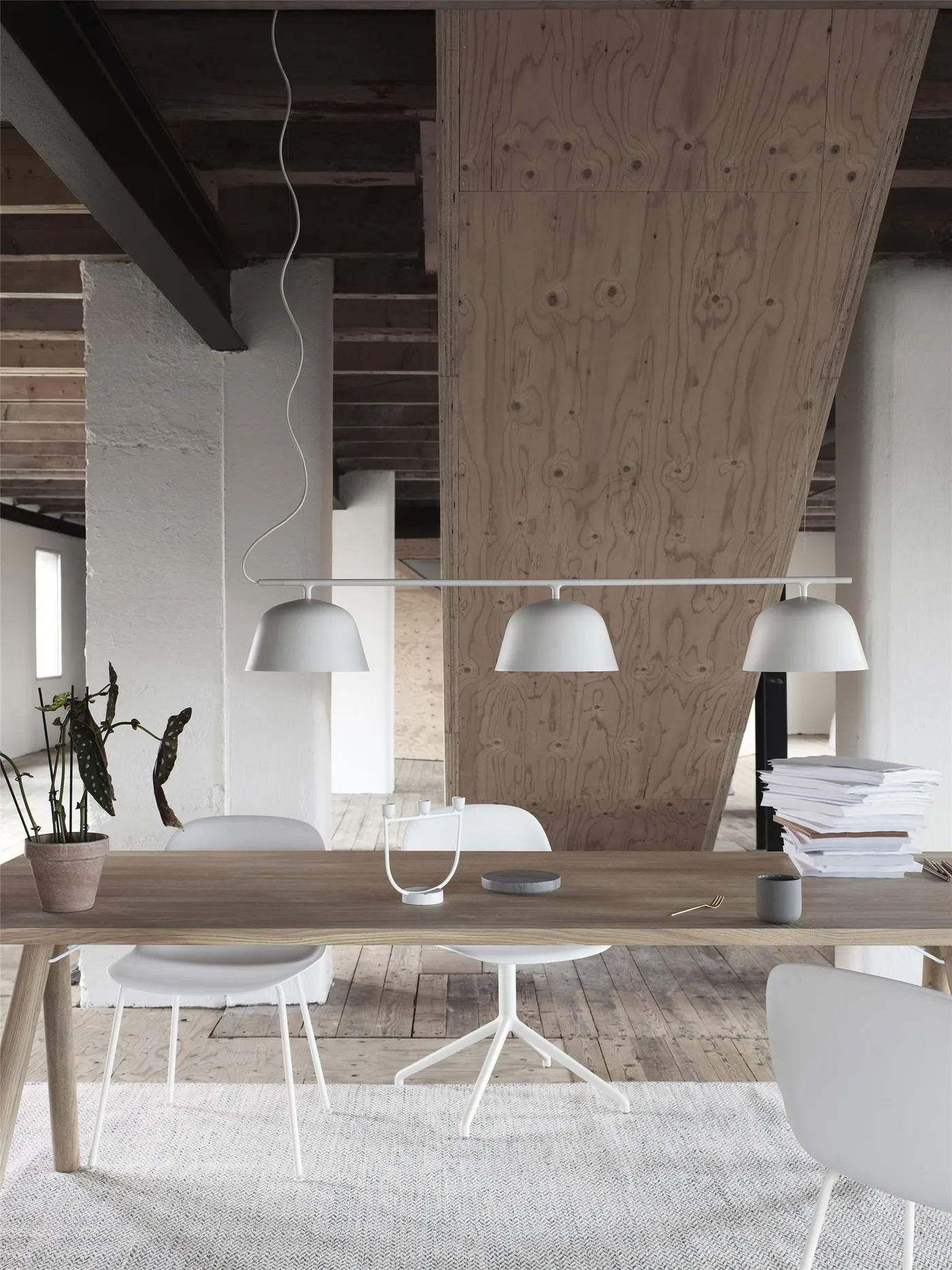 White chairs surround a wooden worktable above a gray textured rug beneath three white pendant shades and exposed roof framing