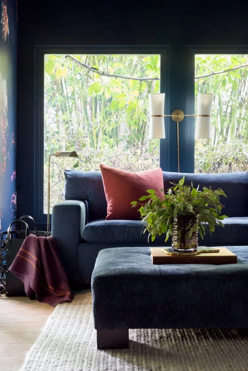 Living room detail with deep blue sofa and ottoman, dark walls, large garden-facing windows, pink cushion and throw, brass-colored light and leafy arrangement.