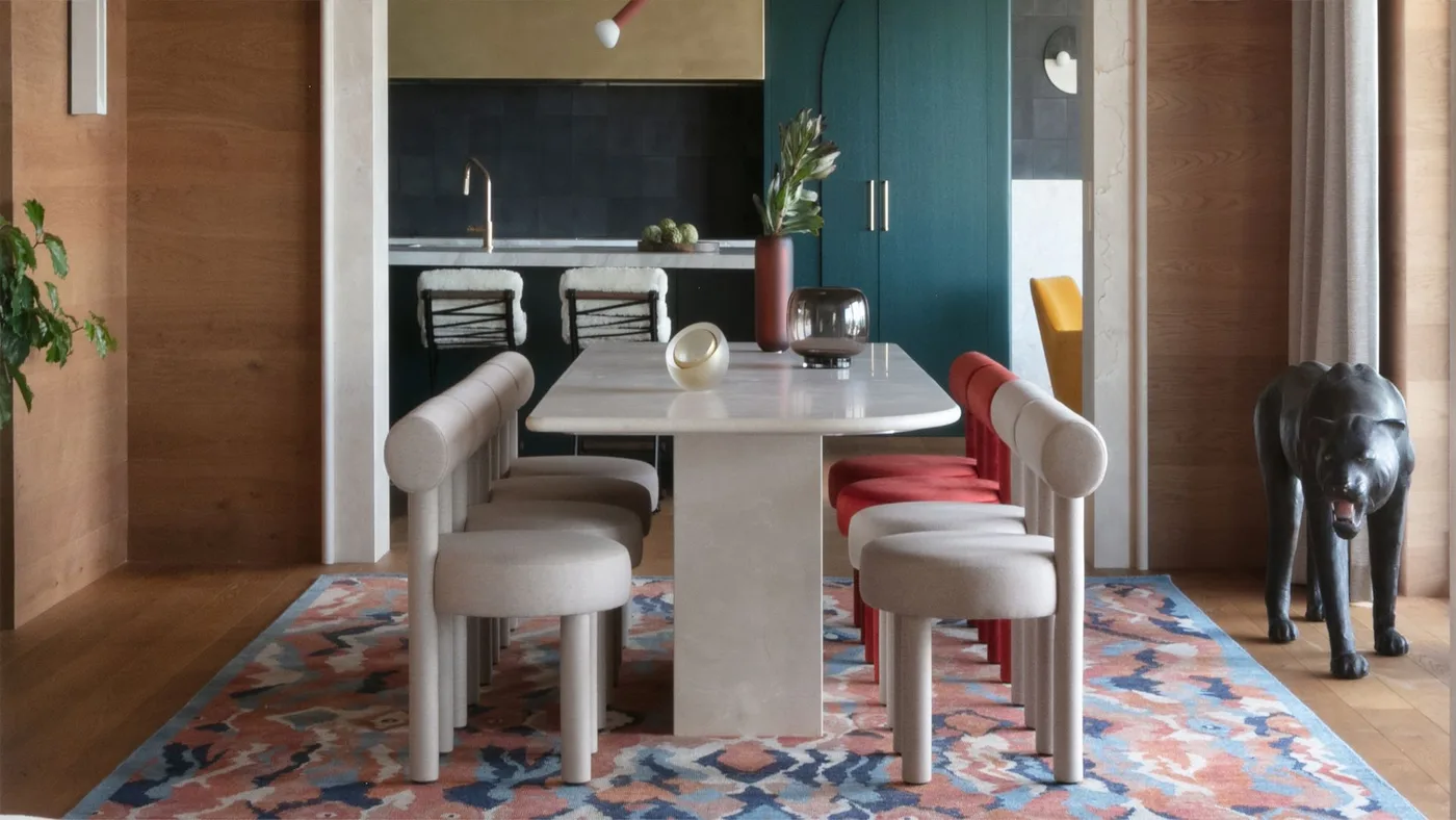 Dining area with a white block-leg table, rounded pale chairs, one red seat, patterned pink-blue rug and warm wood wall panels