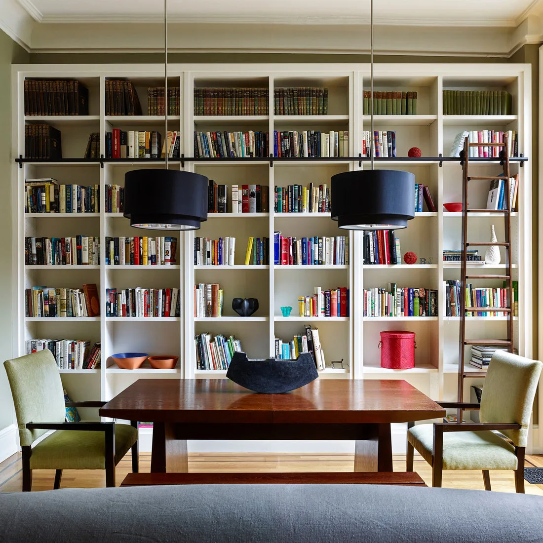 Wood dining table with green armchairs in front of tall white bookshelves and two black drum pendants