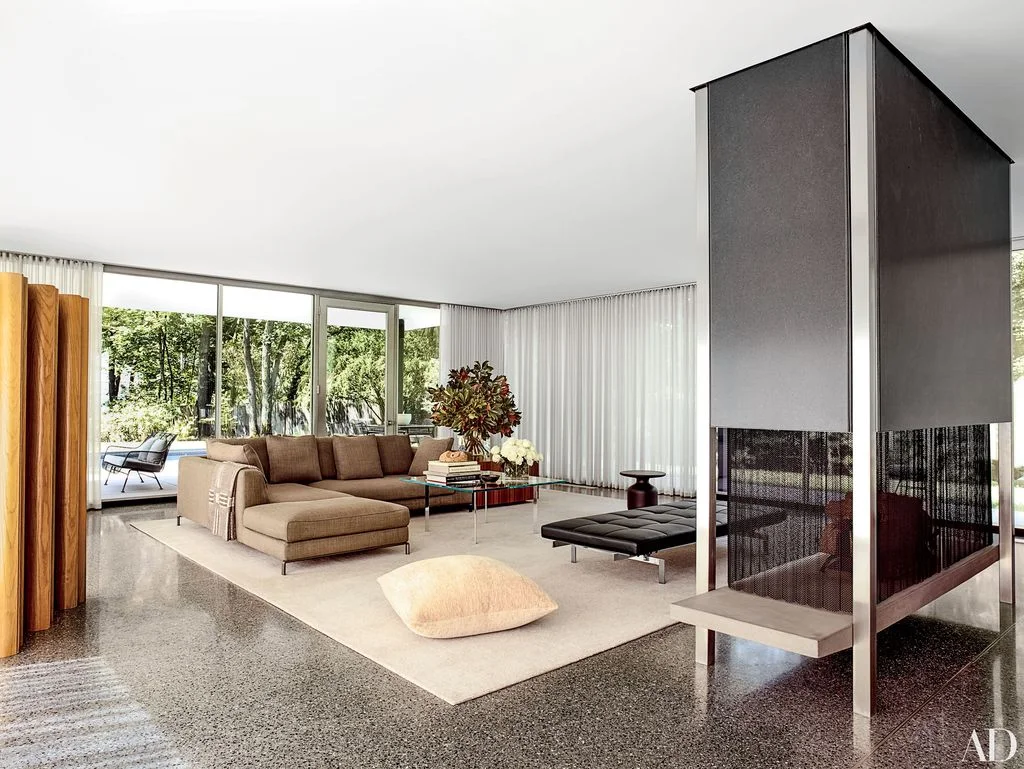 A tall dark freestanding fireplace with screened lower sections stands beside a pale slab-like base, brown sectional seating, broad windows and light curtains