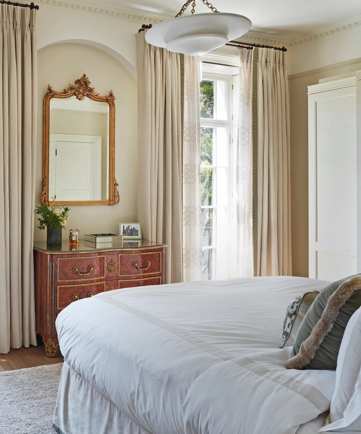 Pale bedroom with long cream curtains, warm red chest, ornate mirror and white bedding beneath a broad hanging light.