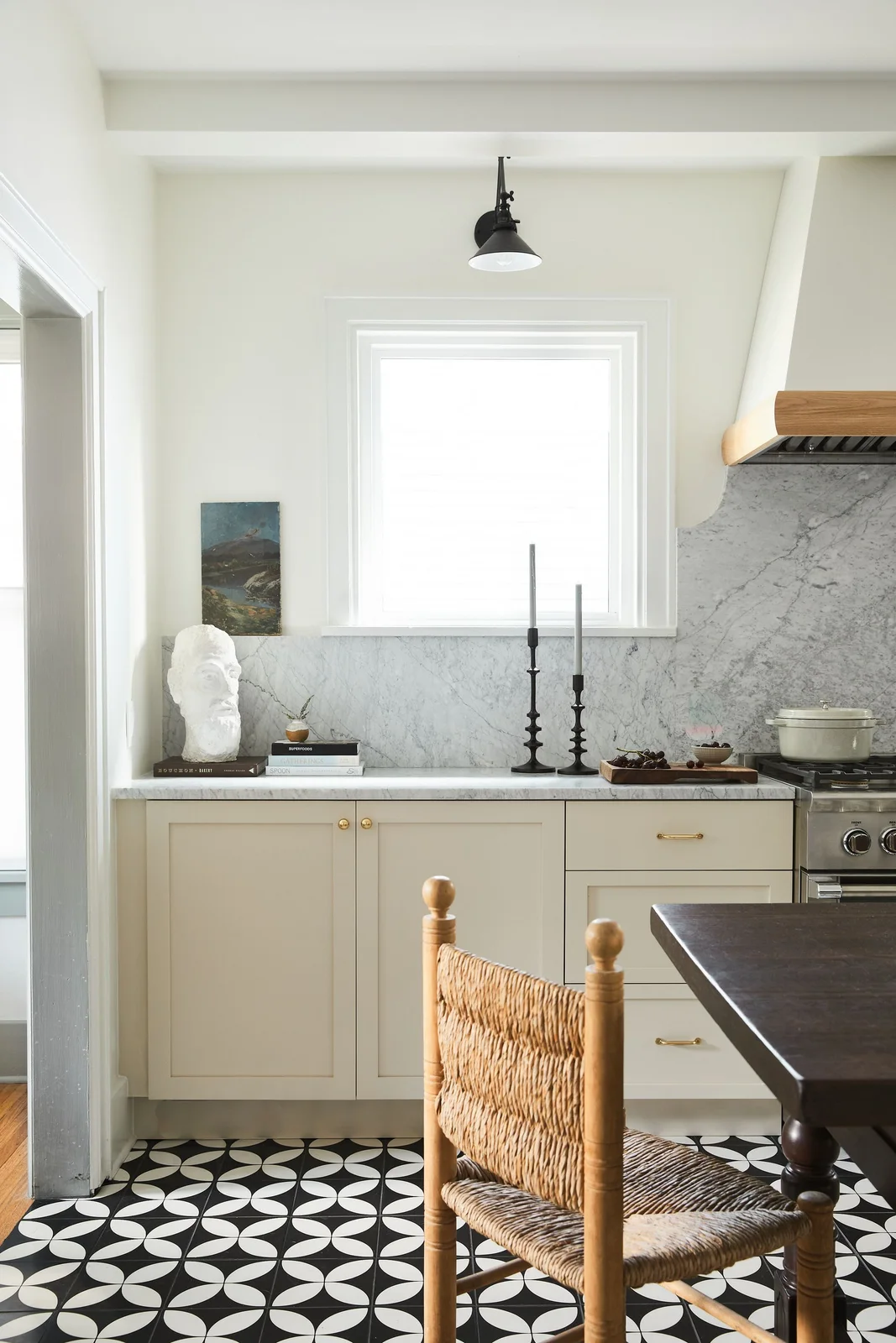 Light beige lower cabinets and a veined backsplash sit beside a black-and-white patterned floor and a woven wooden dining chair