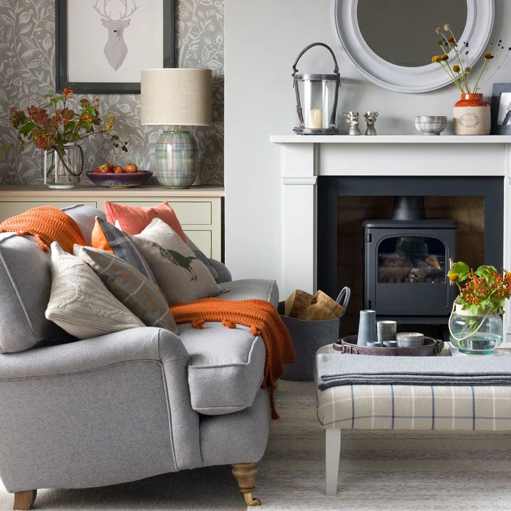 Gray sitting room with stove, round mirror, orange throw and flowers beside upholstered footstool