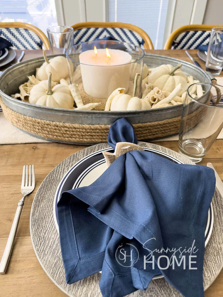 A plain navy napkin rests on a white plate with navy rings above a round charger, with white pumpkins and a tray behind it