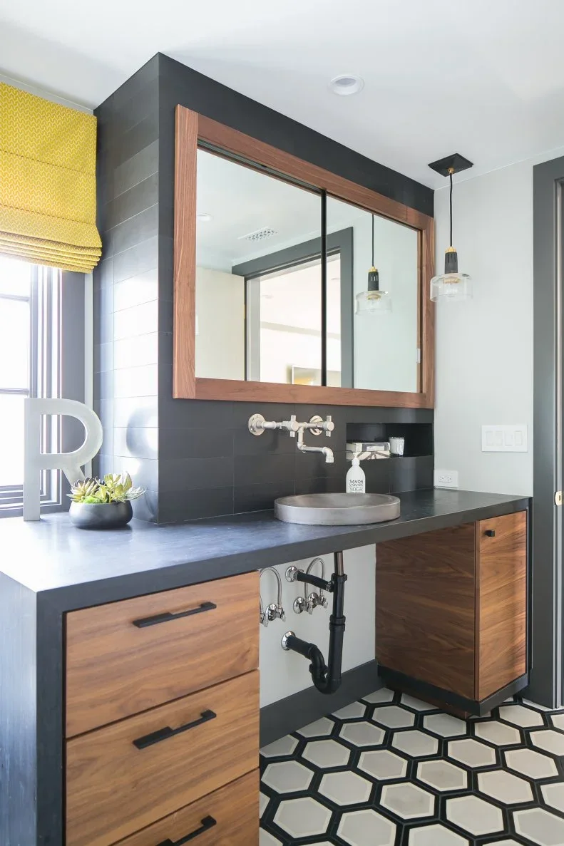 A small round pale basin sits on a dark counter between wood drawer units beneath a large wood-framed mirror against dark wall tiles beside a yellow window shade
