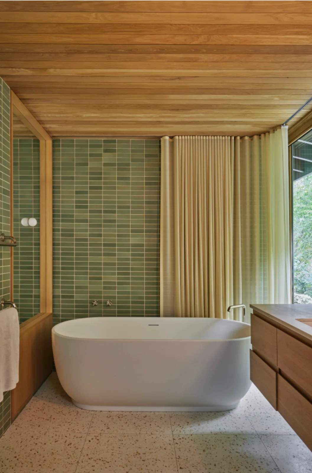 Green grid bathroom tiles sit beside a white freestanding bath beneath a wood ceiling with pale curtains, a speckled floor and wood vanity