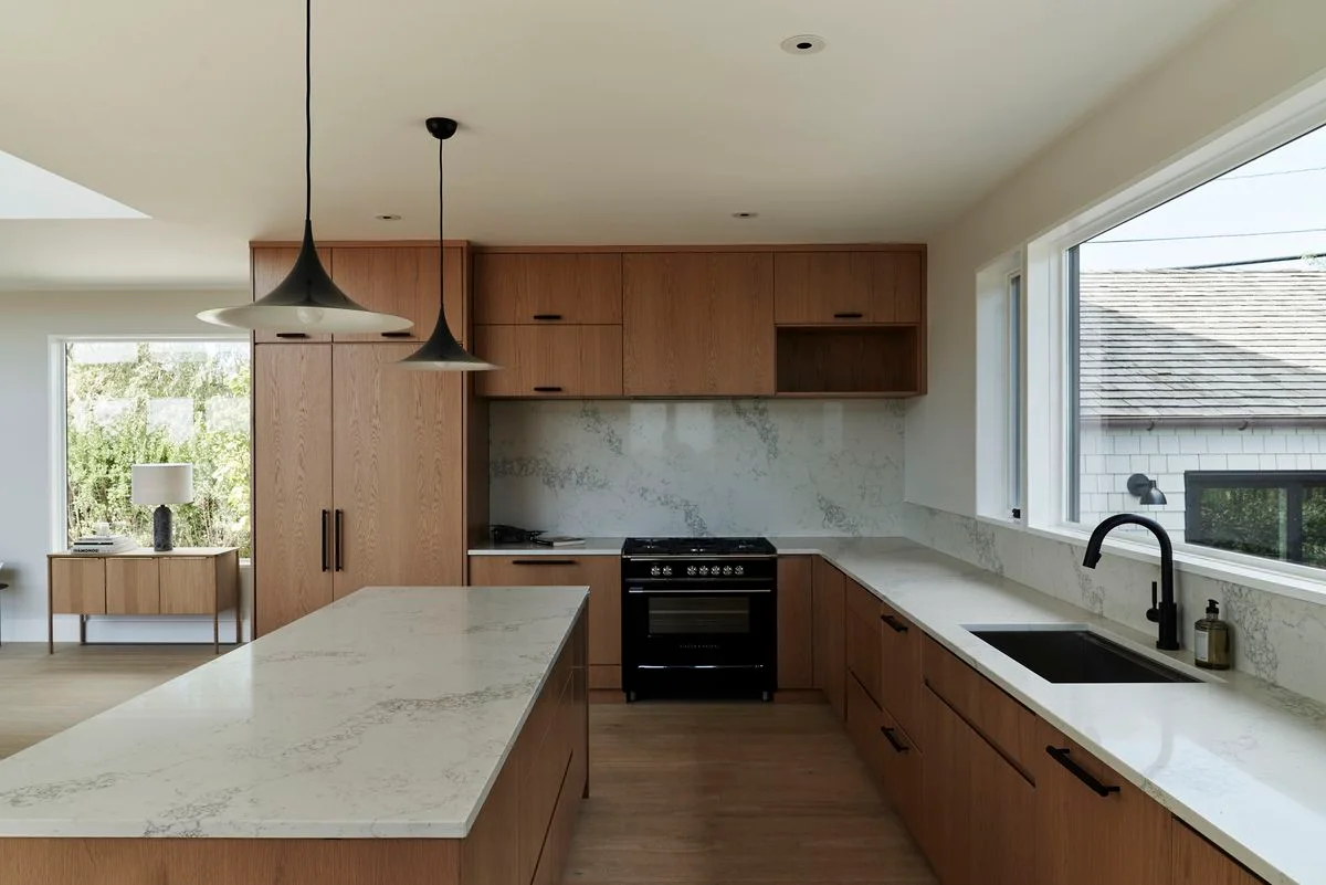 Wooden cabinets surround pale veined counters beside a wide window, black sink and tap, dark cooker and two dark pendant shades
