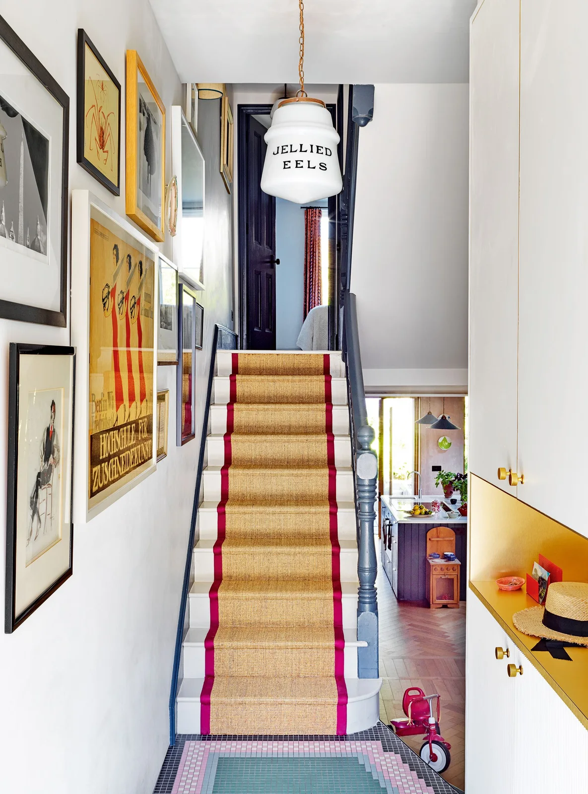 Assorted framed artworks follow a staircase beside a red-edged runner, dark blue-gray railing and a yellow recessed storage area