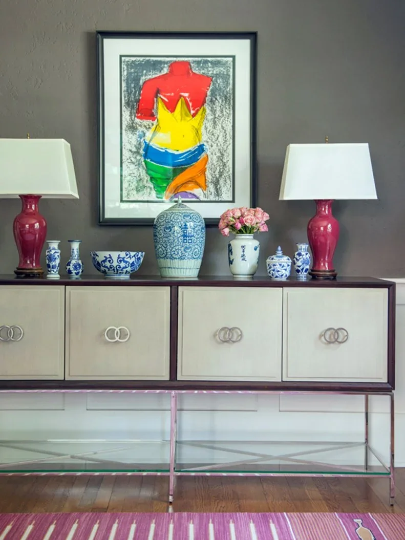 A low sideboard with pale door panels and loop-shaped pulls sits in a darker frame below paired pink lamps and colorful framed artwork