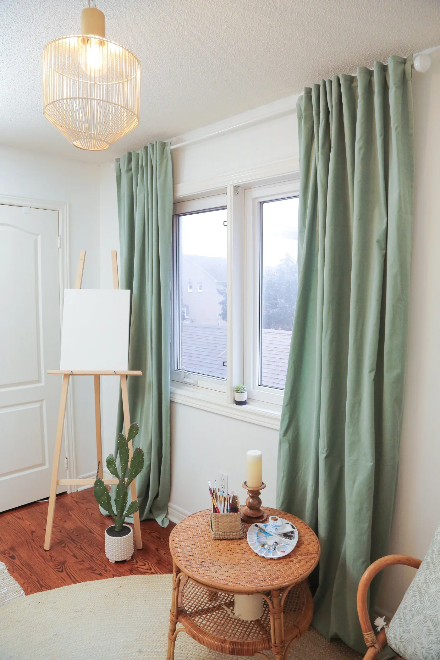 Student bedroom corner with wooden easel, pale green curtains, tall potted cactus and woven round table beside a window.