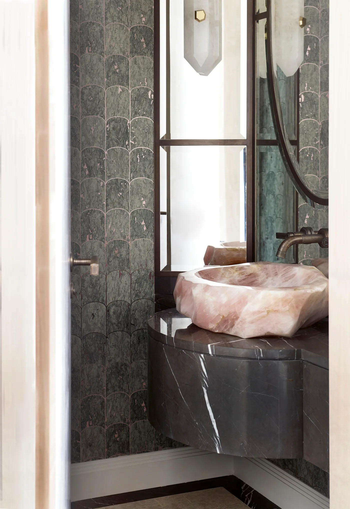 Powder room detail with green scalloped tiles, pink sculptural basin on dark counter, narrow tall mirror, black-framed window and small metallic wall light.