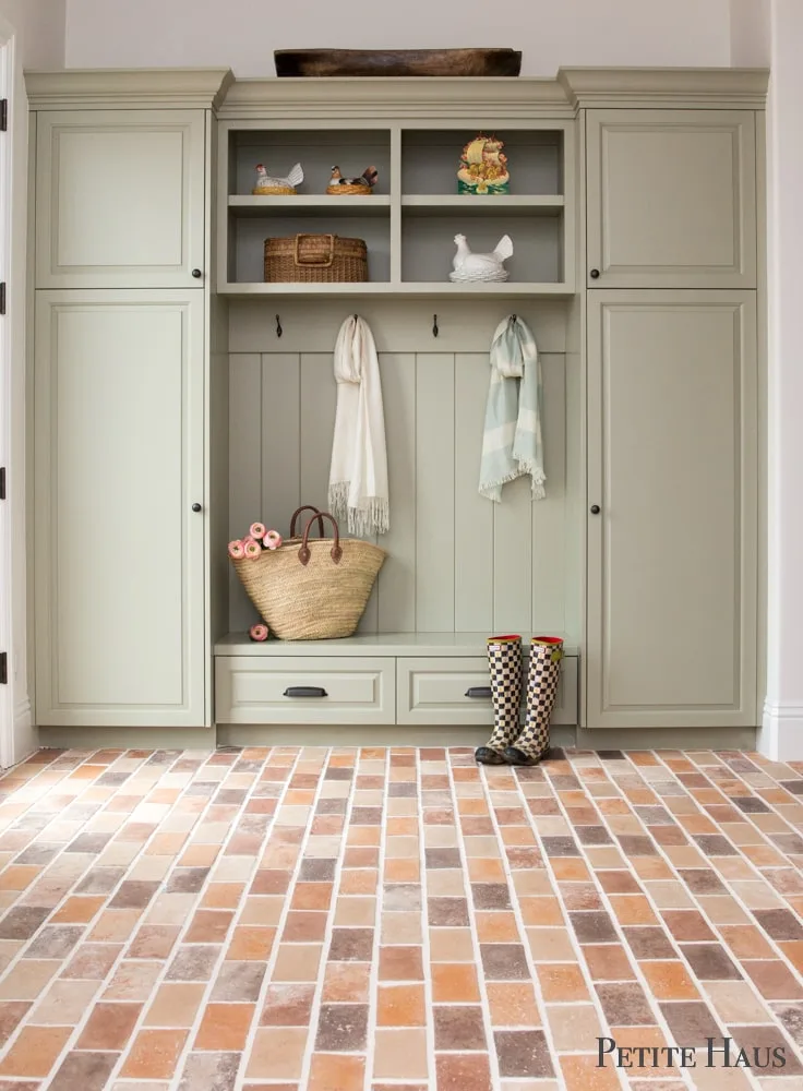 Pale green mudroom cabinet with tall side cupboards, open shelves, hooks and a red brick-toned floor.