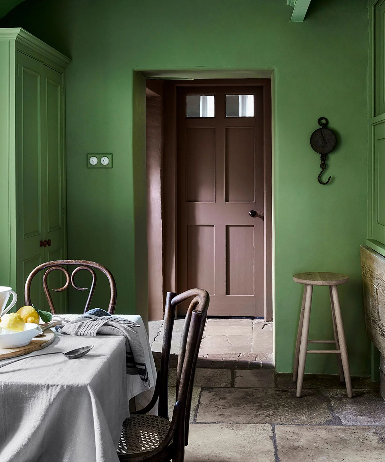 Green walls and painted storage surround a dining table beside dark curved chairs, a brown door and simple wooden stool