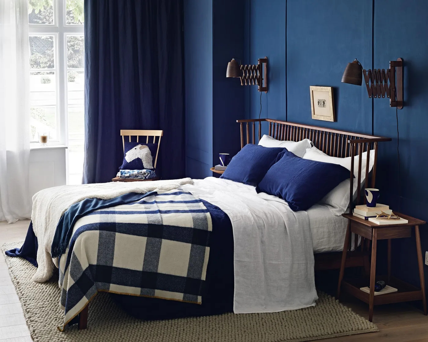 Navy bedroom wall and curtain with a wooden spindle bed, checked blue blanket, white bedding, and extending wall lights.