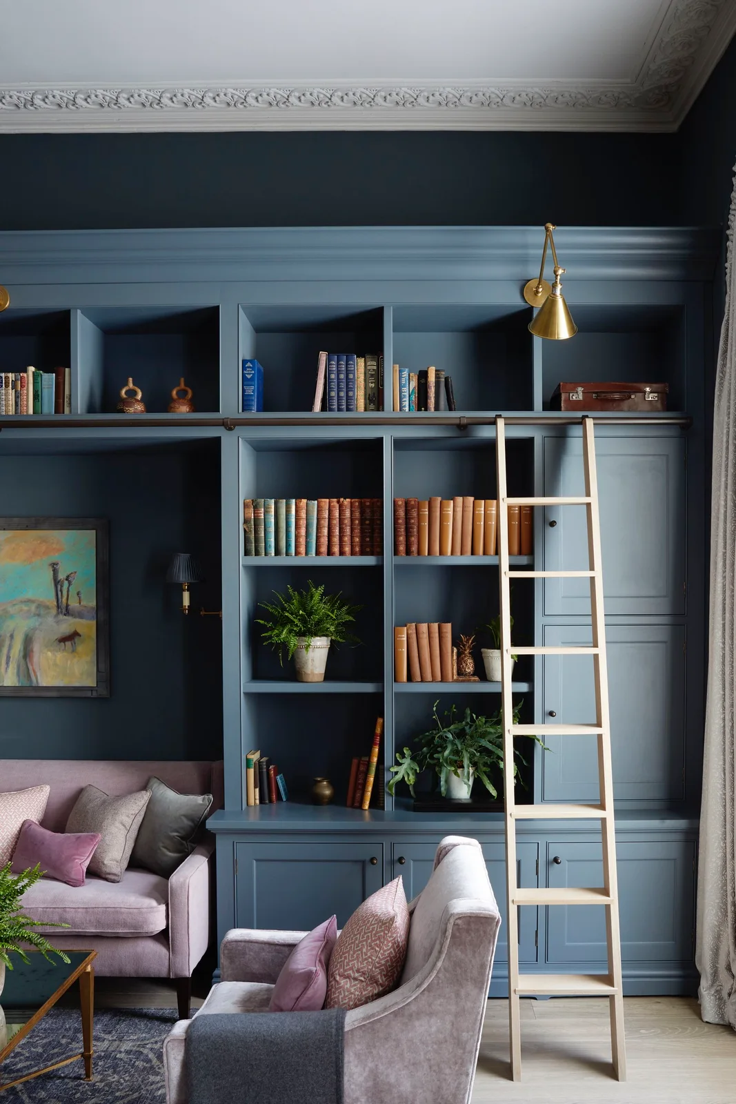 Deep blue shelving with closed lower cupboards surrounds books and small objects beside pale reading seats and a wooden library ladder