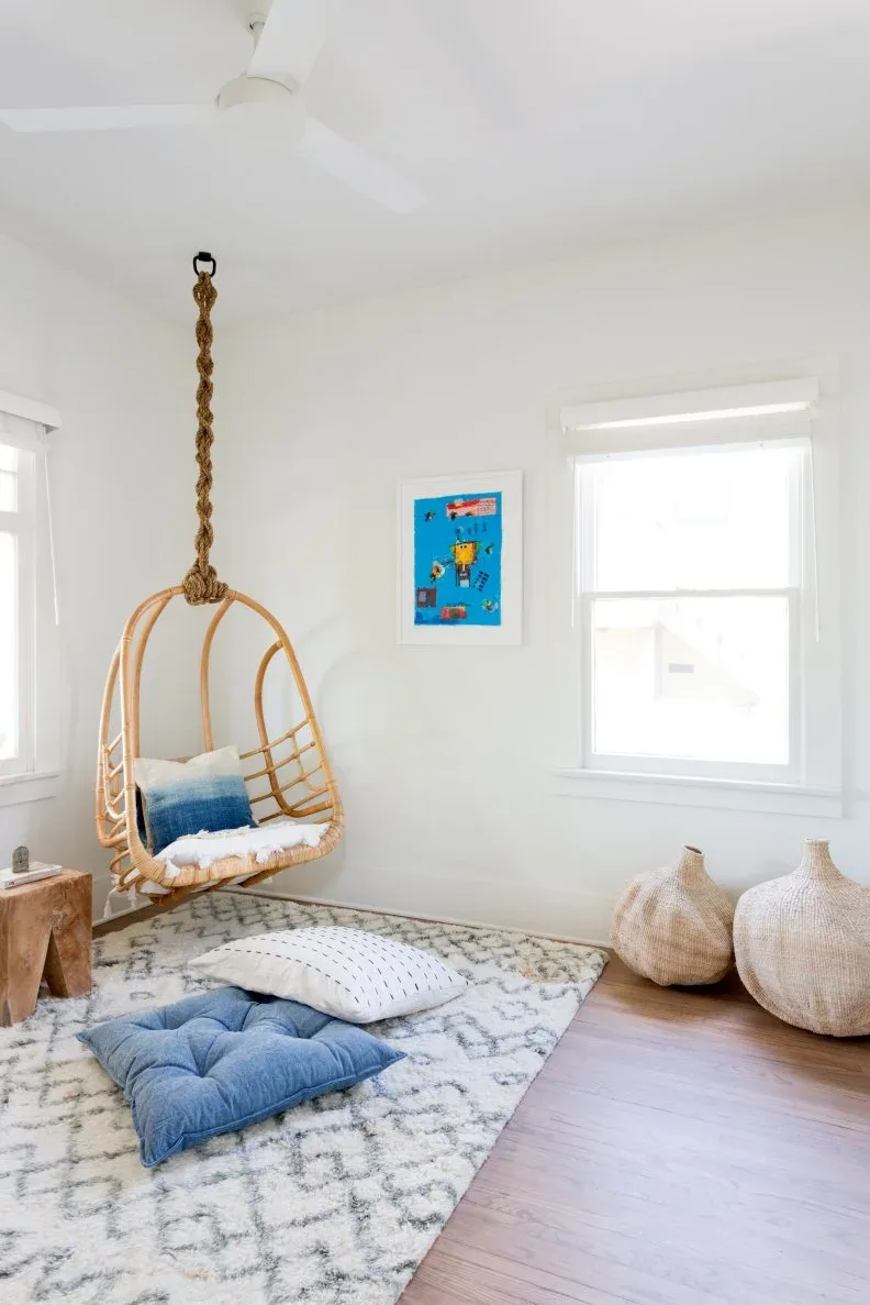 Pale sitting corner with woven hanging chair, light cushions, pale patterned rug, blue floor cushion and two large pale woven vessels.