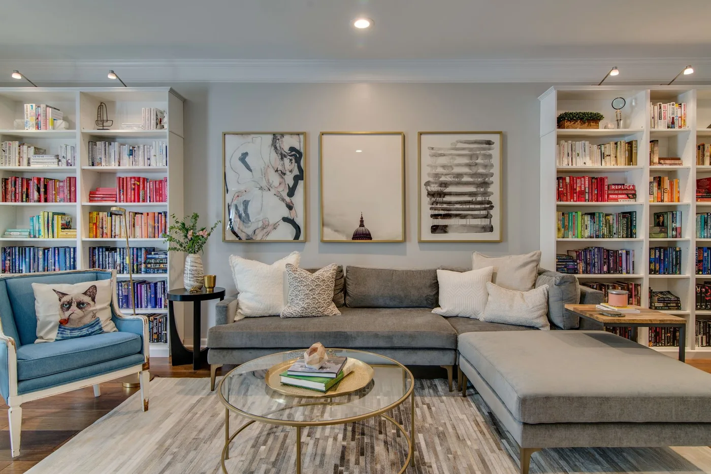 Two white bookcases stand on either side of a gray sectional beneath framed artwork beside a blue chair and round glass-topped table