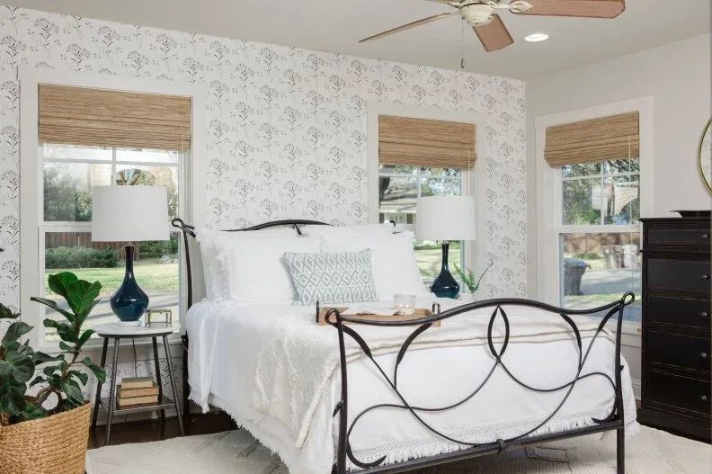 Pale botanical bedroom wallpaper with warm woven shades, a dark curved bed frame and a leafy plant