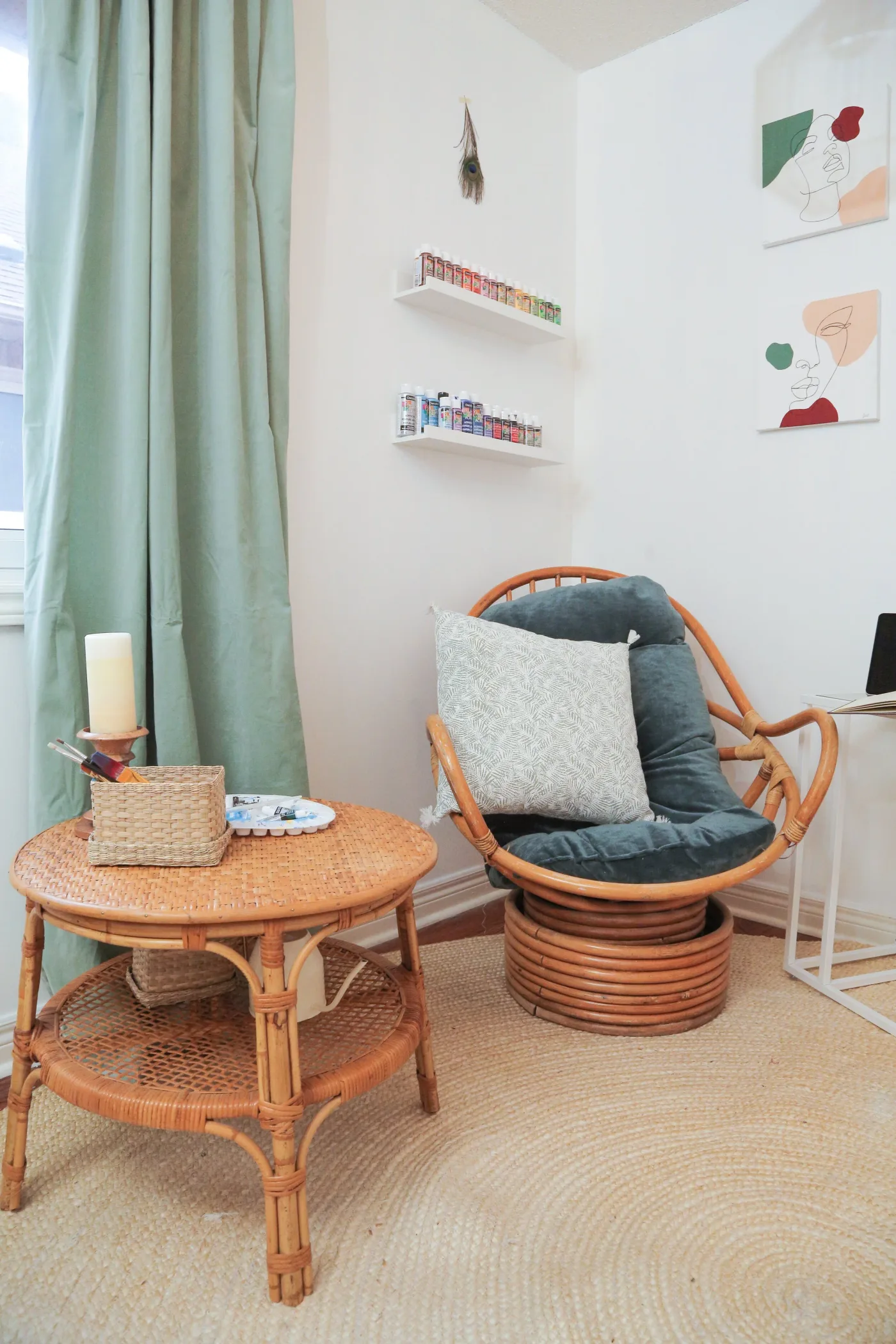 Rounded wooden chair with dark green cushion beside a woven round table, pale green curtain and narrow art-supply shelves.