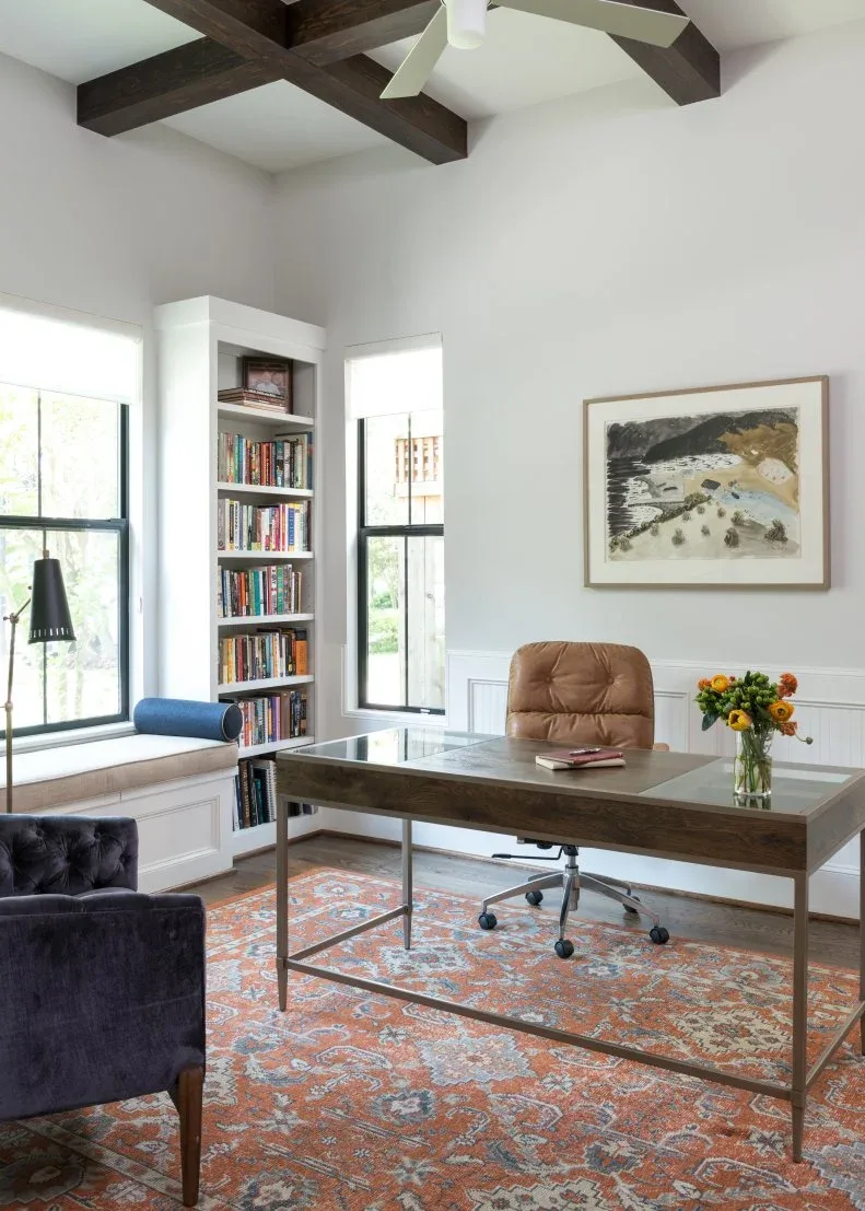 Home office with open rectangular desk, tan rolling chair, orange-and-blue patterned rug, tall bookcase between windows, pale walls and framed artwork.