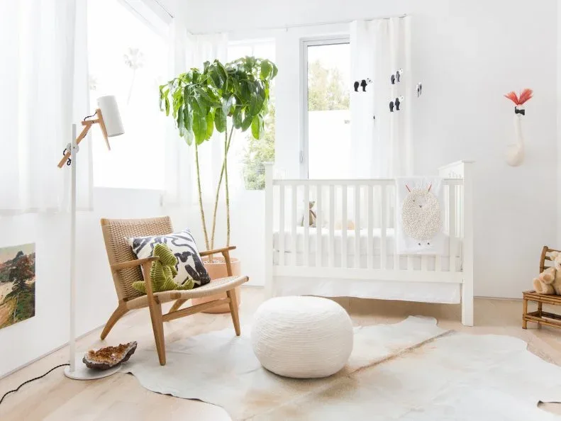 A white crib stands beside a woven wooden chair, pale round pouf, floor lamp and large plant in a white nursery