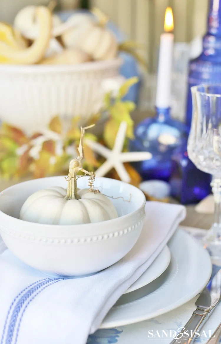 A small white pumpkin with a gold-colored stem sits in a white bowl above layered plates beside a blue candle bottle and a small deer ornament
