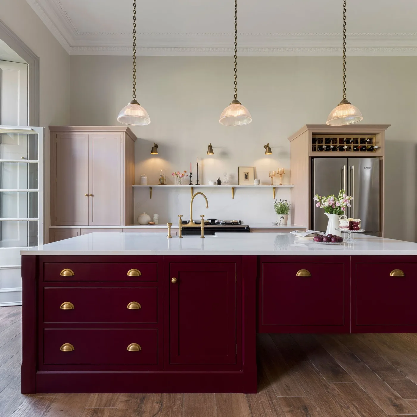 Kitchen with a burgundy framed island, pale worktop, gold-colored cup handles and three pendant lights above.