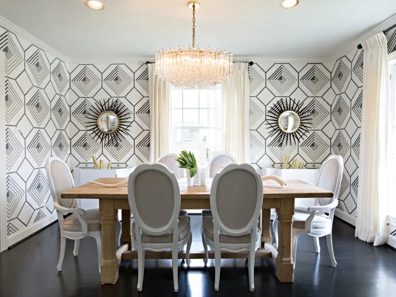 Traditional pale round-back chairs beside a wooden rectangular table, geometric wallpaper, two sunburst mirrors and a rod chandelier.