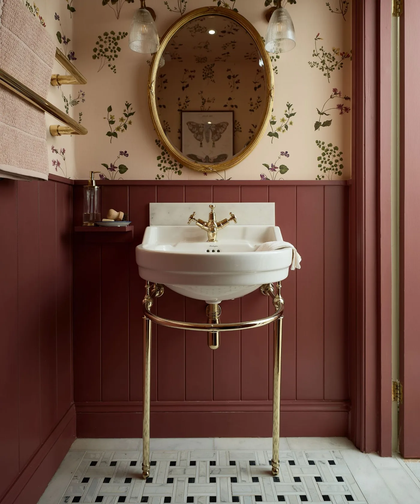 Powder room with small botanical wallpaper, burgundy lower wall paneling, oval gold-framed mirror, white console basin, pale floor and glass-shaded lights.