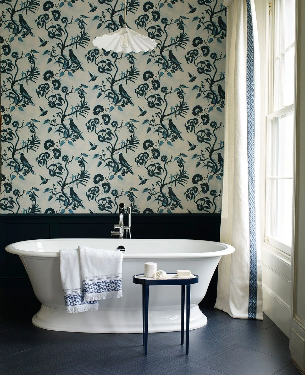 Bathroom with large dark blue floral wallpaper, dark lower paneling, white freestanding bath and a small dark blue side table.