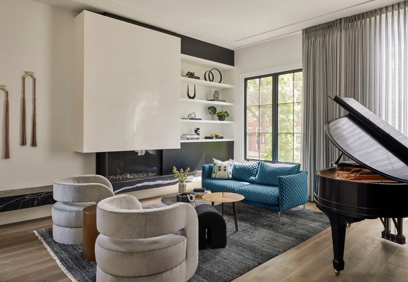 A pale projecting fireplace box sits above a long dark firebox and veined hearth beside curved gray chairs, a teal sofa and piano
